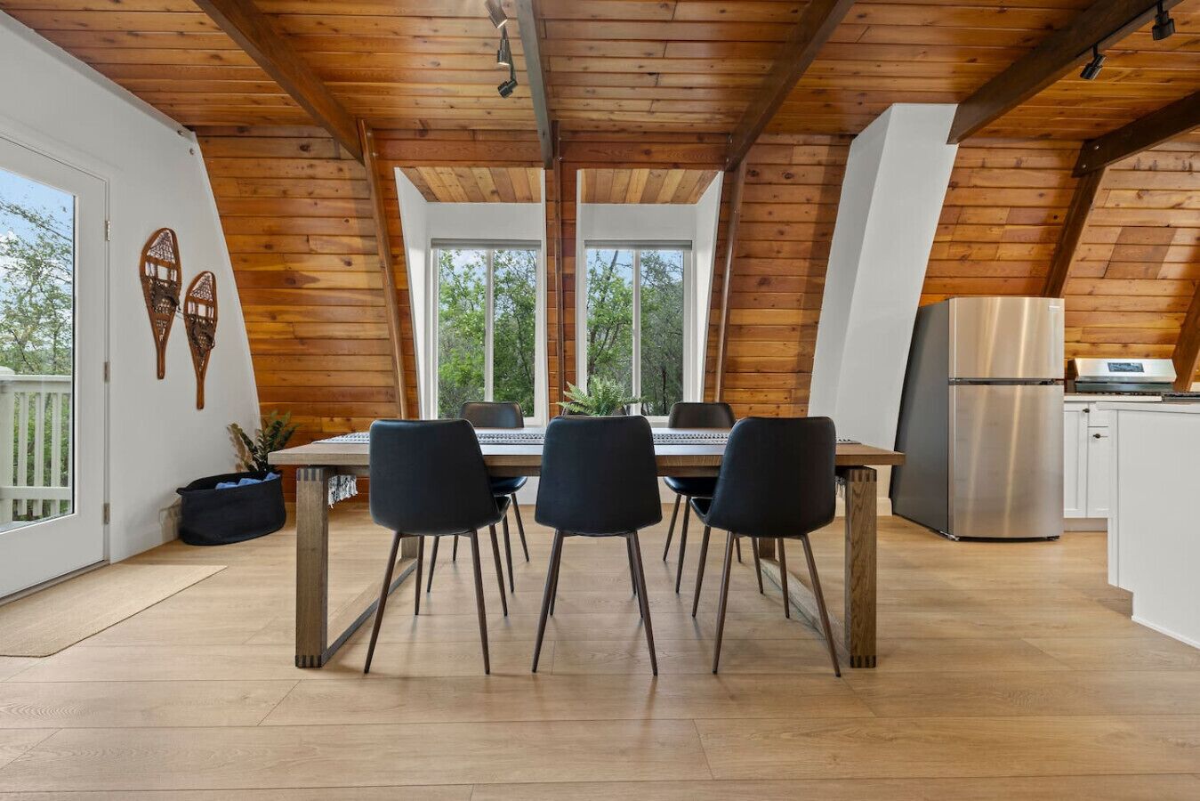 A modern kitchen with wooden beams, a dining table with four chairs, a refrigerator, and large windows.