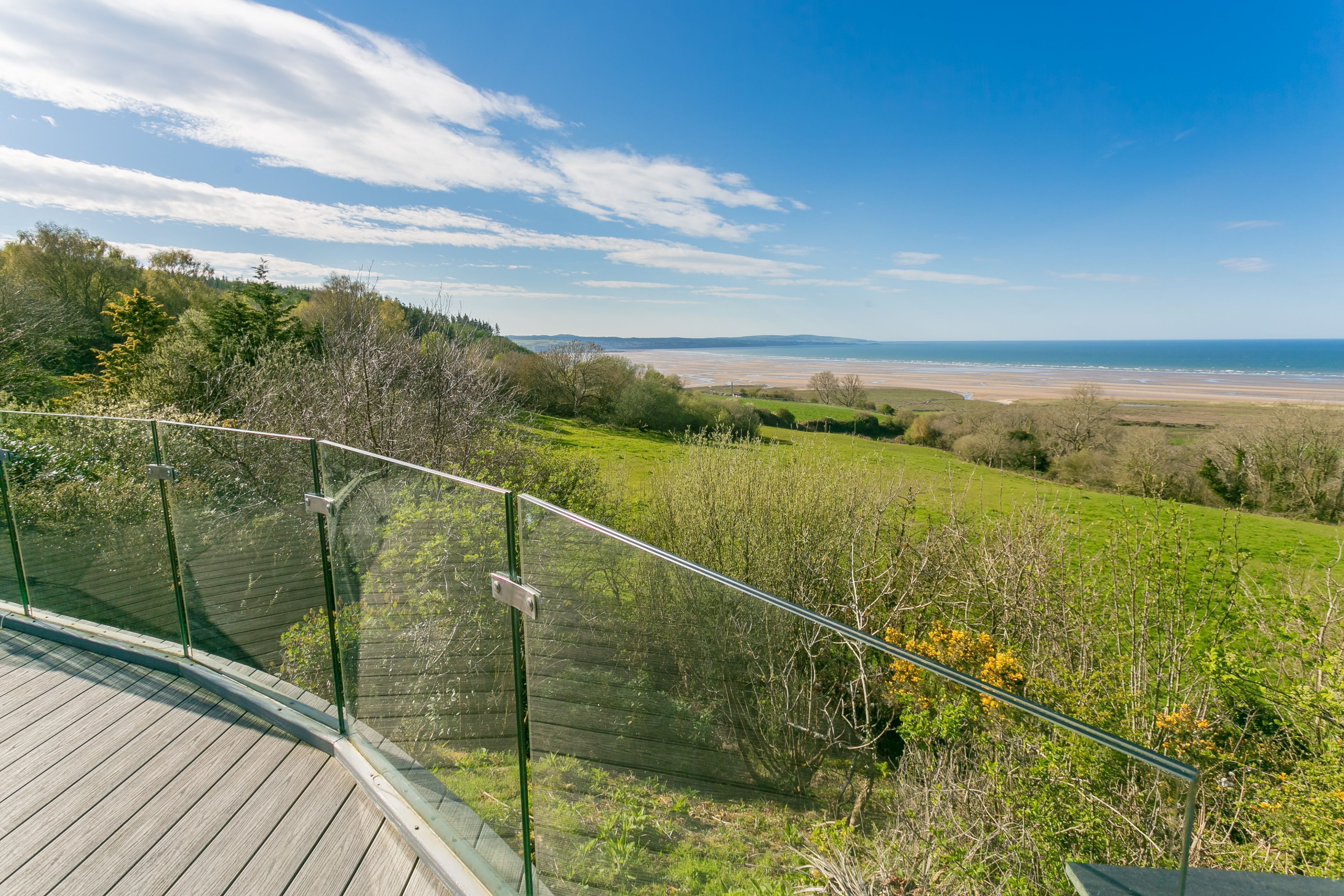 Une terrasse en bois dotée d’une rambarde en verre, offrant une vue sur un paysage côtier avec des champs verdoyants et une plage.