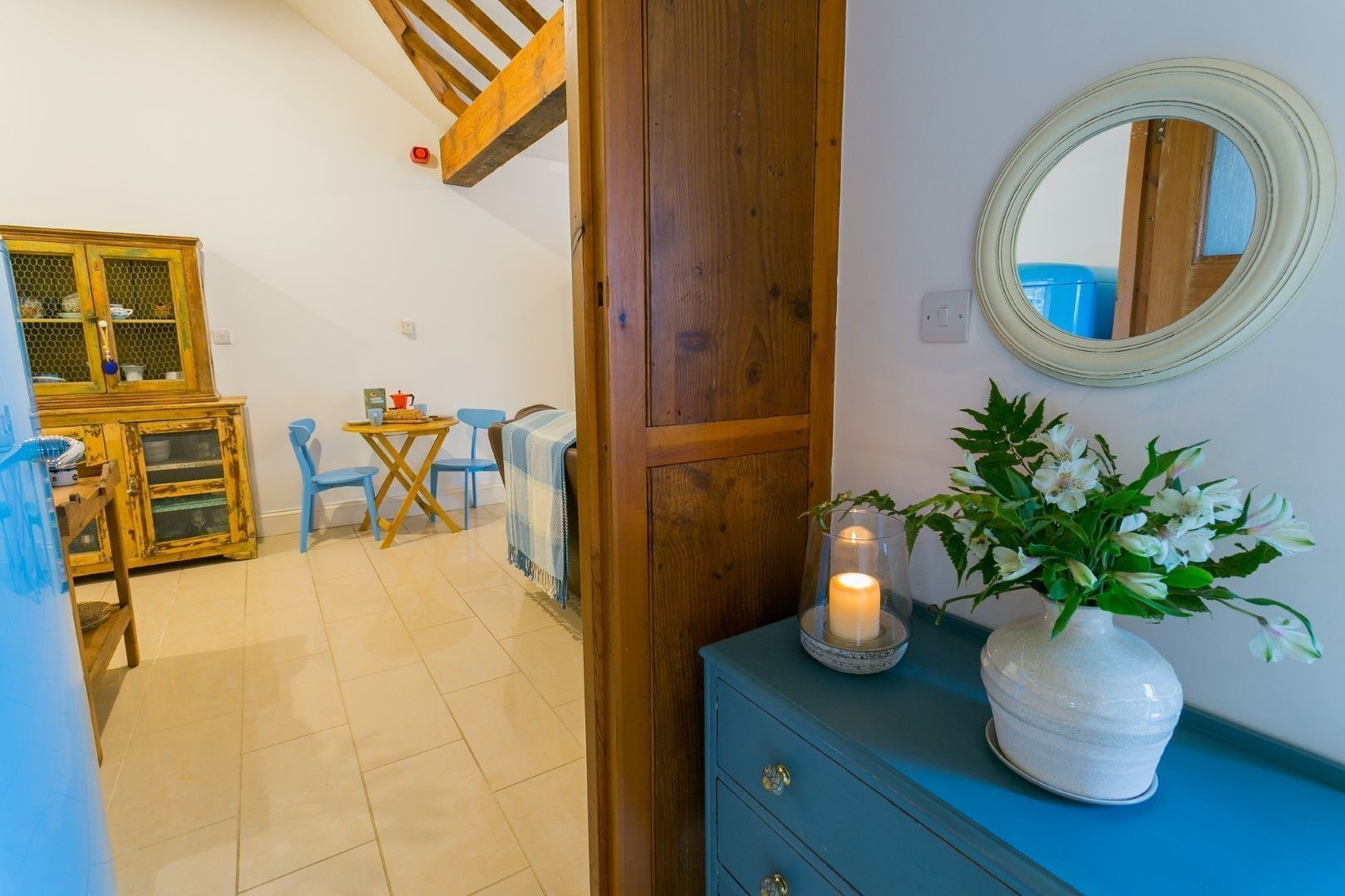 A room with a wooden door, a blue dresser, a mirror, a vase with flowers, a lit candle, and a dining area with a table and chairs.