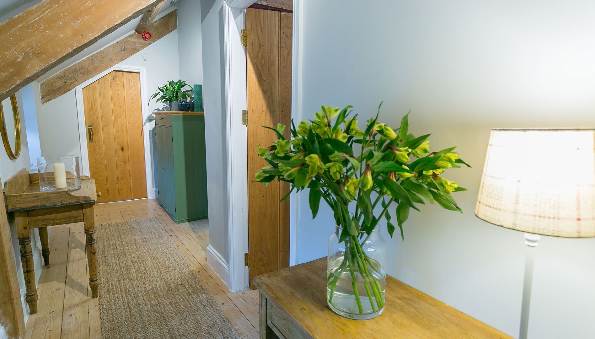 A hallway with a wooden floor, a rug, a wooden bench, a vase with flowers, a lamp, and a door leading to another room.