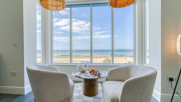 A room with two white chairs facing a large window overlooking a beach.