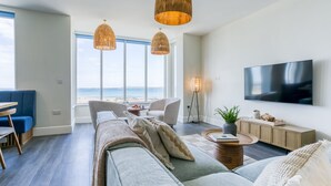A modern living room with a large window overlooking the beach, a sofa, a TV, and a dining area.