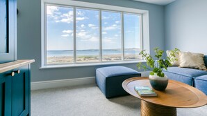 A room with a large window overlooking a beach, a blue sofa, a wooden coffee table, and a plant.