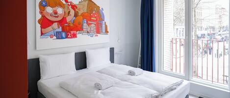 A hotel room with a bed, white bedding, a large window with a view of the city, and a colorful artwork on the wall.