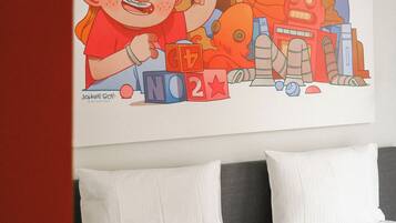 A hotel room with a bed, white pillows, and a colorful artwork depicting a child playing with blocks and robots.