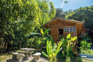 A wooden cabin with a porch, surrounded by lush greenery and a small garden with a table and benches. - Cabaña Ave (Provincia de Guanacaste)