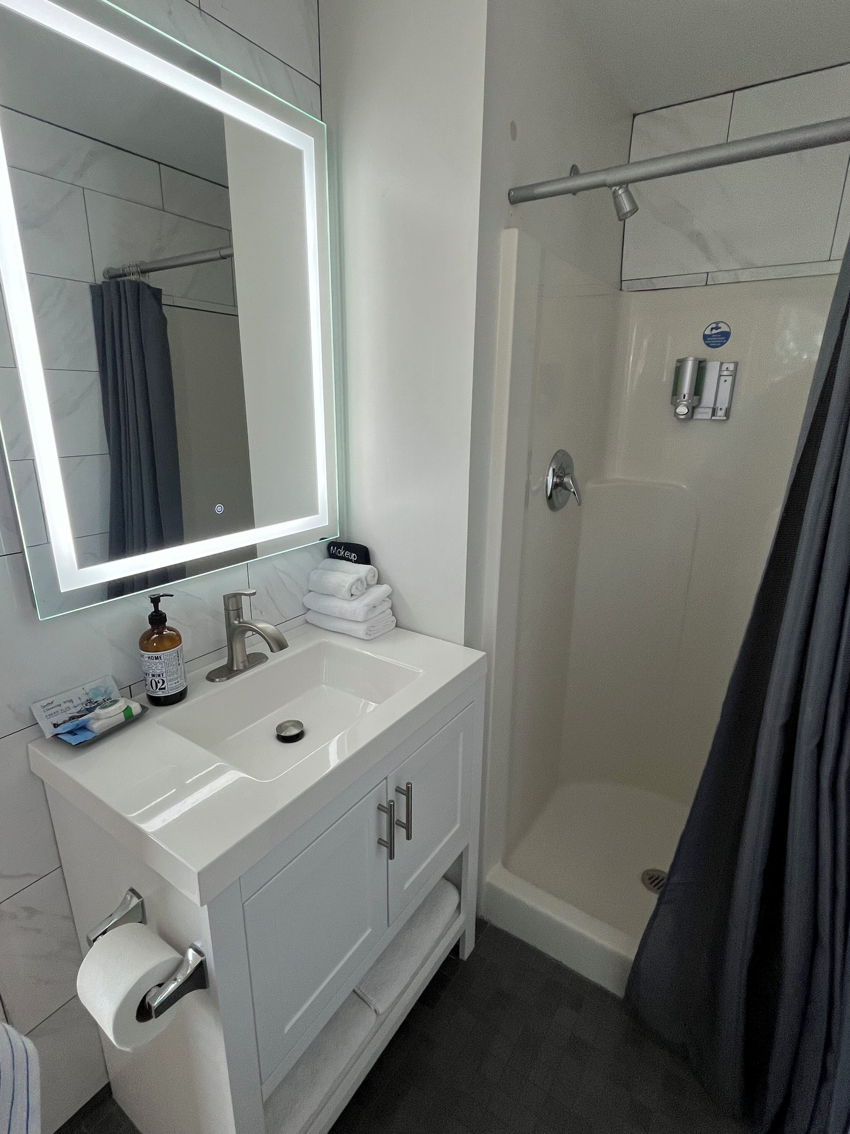 Un bagno con un mobile lavabo bianco, uno specchio con luce integrata, una doccia e un porta asciugamani.