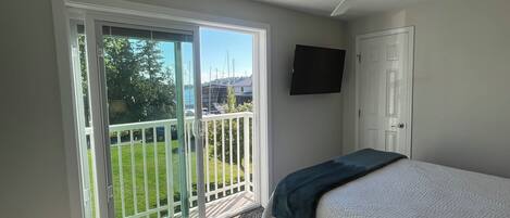 A bedroom with a bed, a ceiling fan, a TV, and a sliding glass door leading to a balcony with a view of the outdoors.