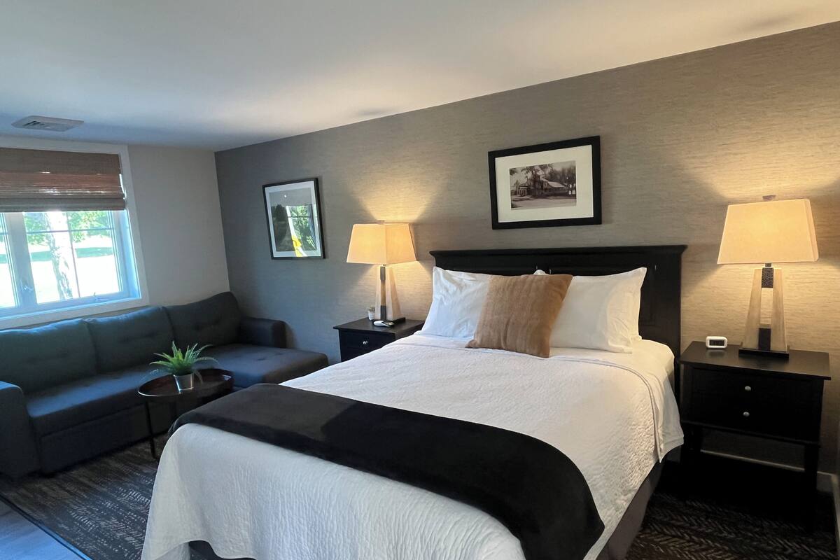 A hotel room with a bed, two bedside tables with lamps, a nightstand, a sofa, and a window with blinds.