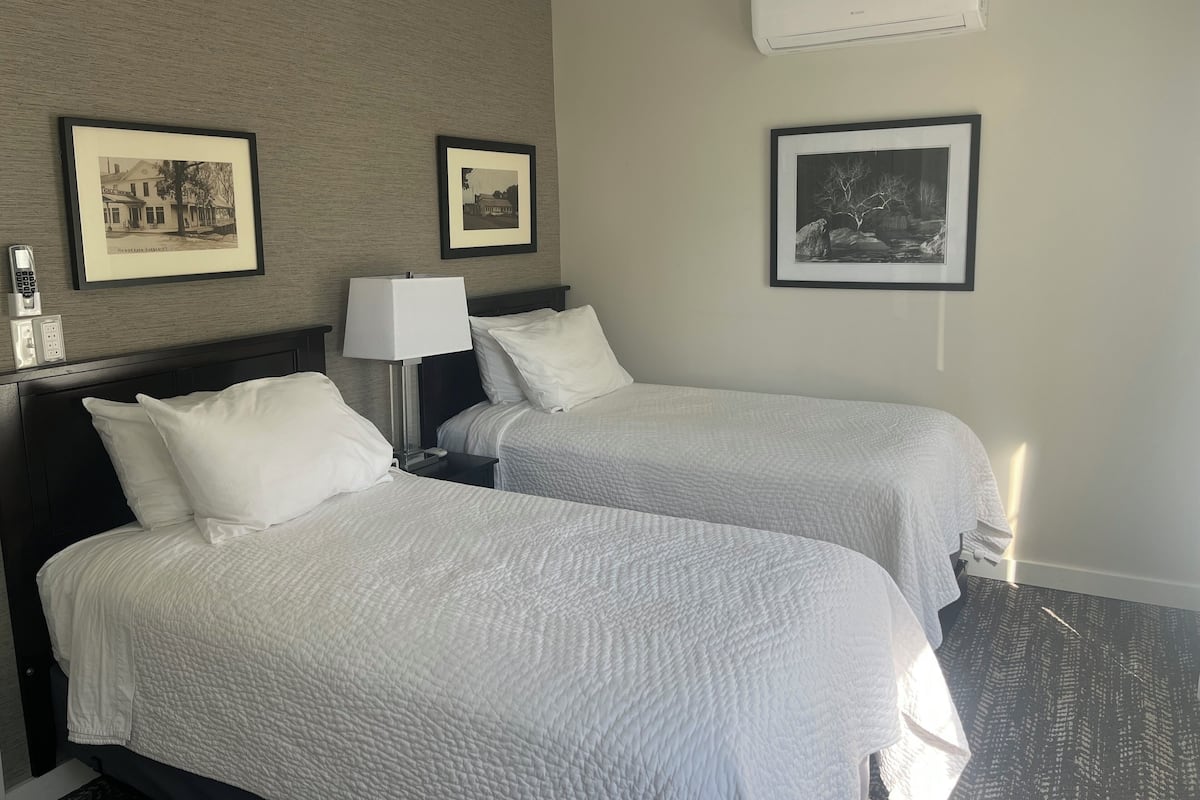 A hotel room with two beds, a nightstand, a lamp, and three framed pictures on the wall.