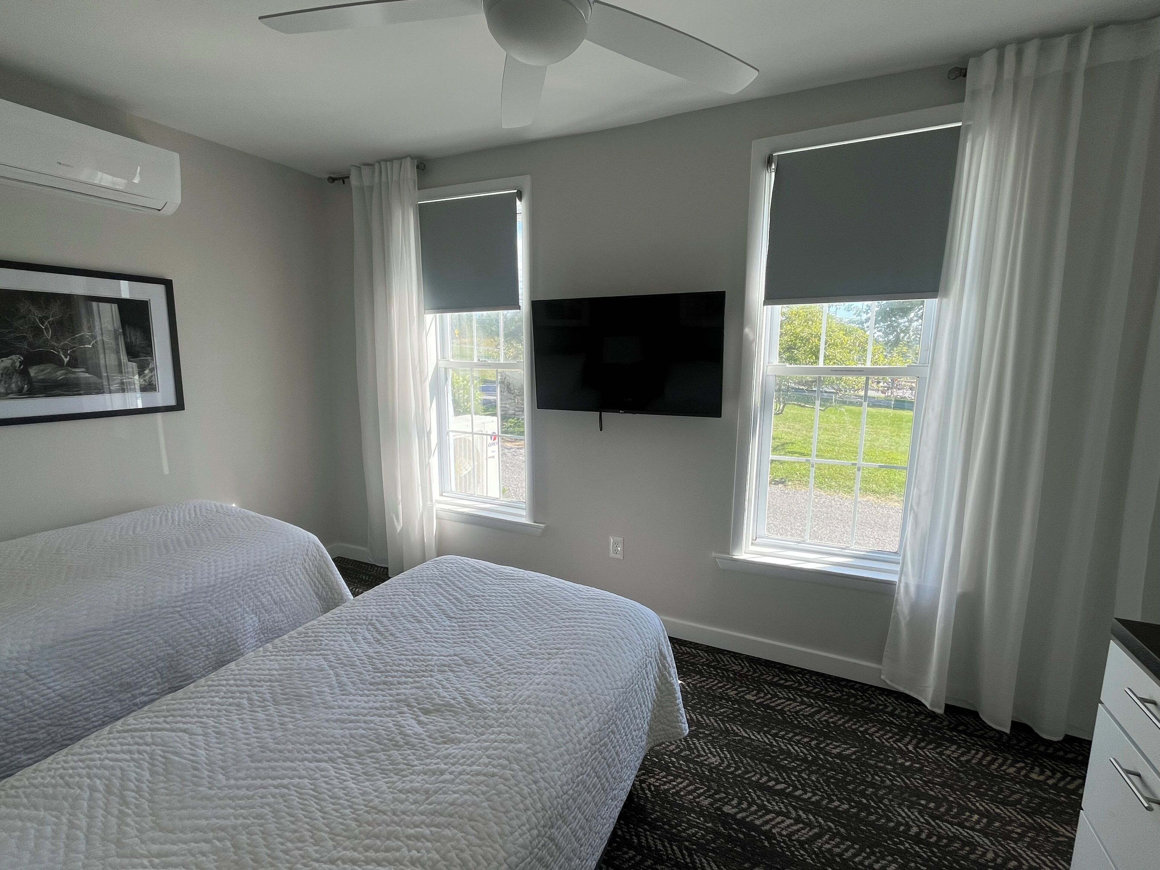 Habitación de hotel con dos camas, ventilador de techo, televisor de pantalla plana montado en la pared y ventanas amplias con cortinas blancas.