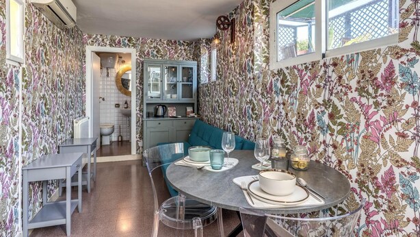 A compact kitchen with a dining area, featuring a round table with clear glass legs, a teal sofa, and a window with a view of the outside. - One Bedroom Apartment (ROMA)