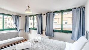 A hotel room with a bed, a sofa, a window with curtains, and a hanging light fixture. - One Bedroom Apartment (ROMA)