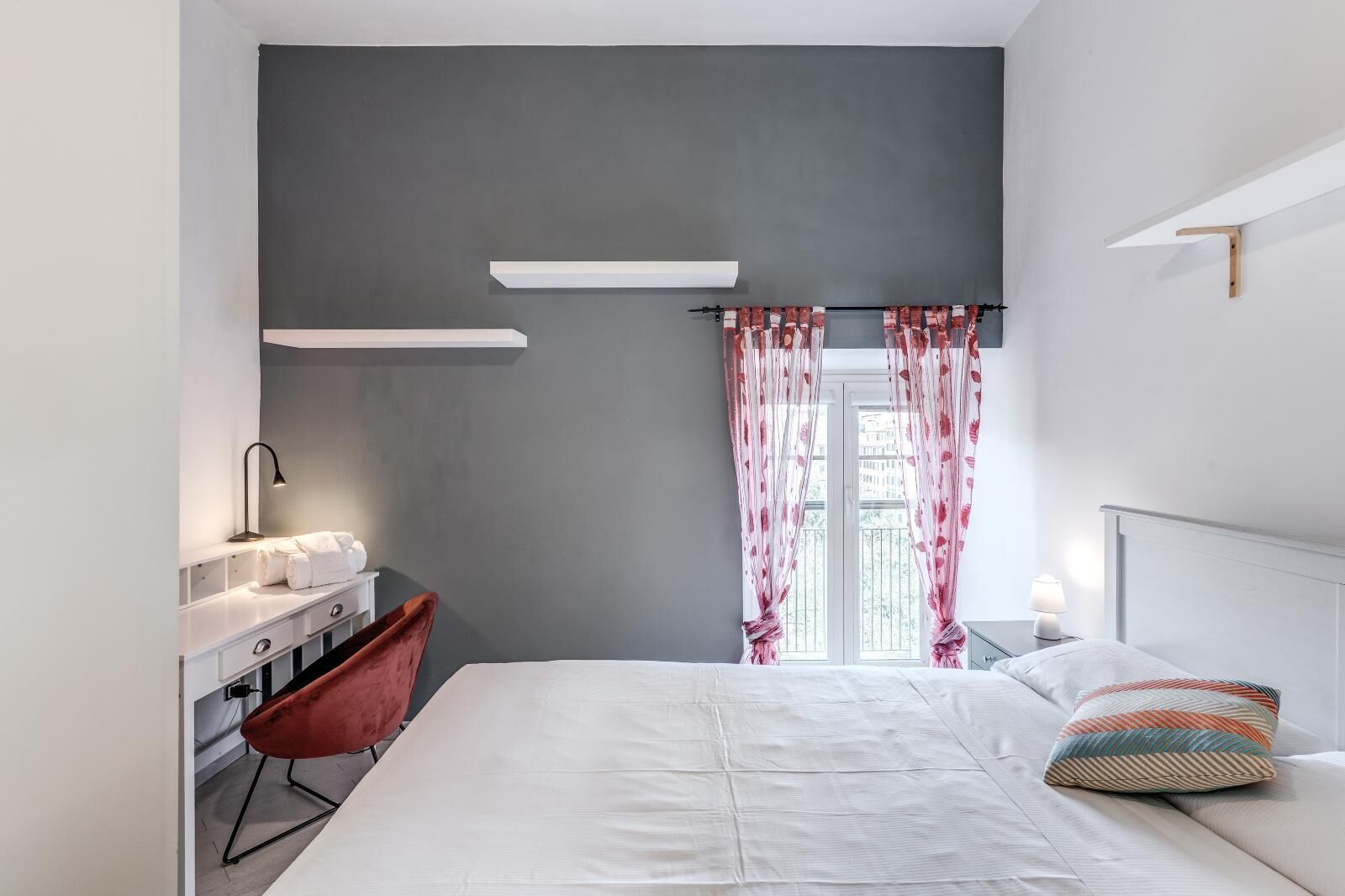 A modern bedroom with a bed, a desk with a chair, a bedside table, and a window with curtains.