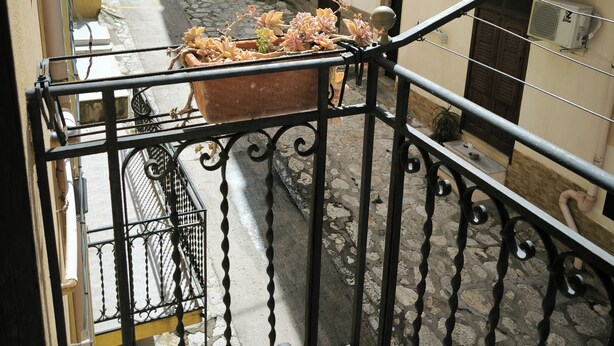A balcony with a wrought iron railing overlooking a narrow street with buildings. - Trinity House (Castellammare del Golfo)