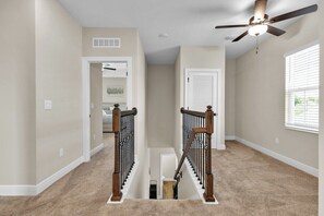 Un escalier recouvert de moquette, avec un ventilateur de plafond, une porte et une fenêtre munie de stores.