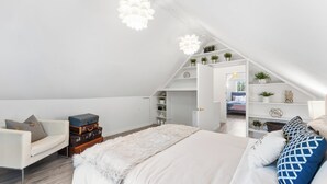 A modern bedroom with a loft-style ceiling, a white sofa, a large bed with patterned pillows, and a white fur throw.