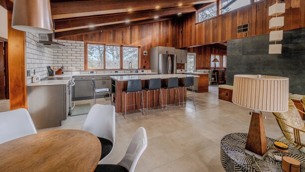 A spacious kitchen with wooden accents, a central island with bar stools, and a dining area with a table and chairs. - Mid-Century Haven with Private Lakes and Endless Views (Erhard)