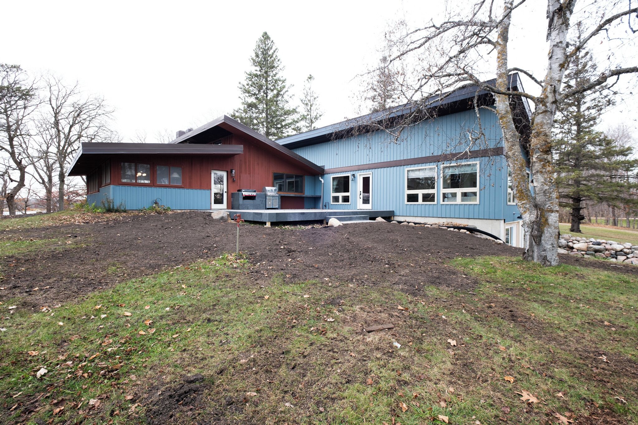 A two-story house with a blue exterior and a brown roof, surrounded by a landscaped yard with trees and a stone wall.