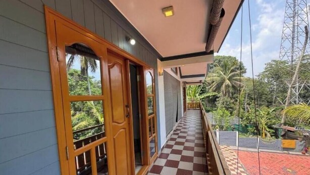A corridor with a checkered floor leading to a wooden door with glass panels, overlooking a lush garden and a red-roofed structure.