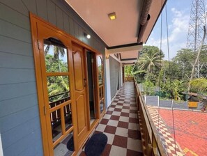 A corridor with a checkered floor leading to a wooden door with glass panels, overlooking a lush garden and a red-roofed structure.