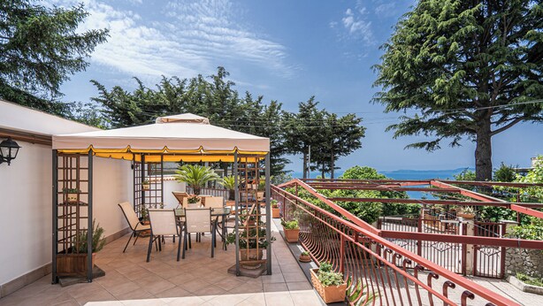 A covered outdoor seating area with tables and chairs, surrounded by potted plants and a red railing.