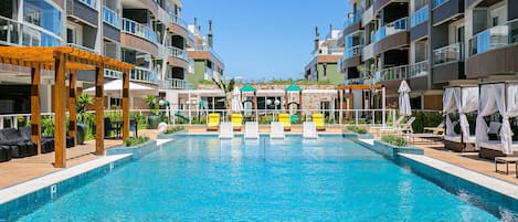 A swimming pool with lounge chairs and umbrellas, surrounded by multi-story buildings with balconies.