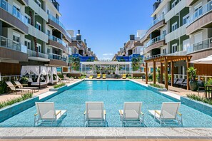 A swimming pool with lounge chairs, a covered seating area, and a view of multi-story buildings. - Seaside Condominium in Campeche-florianopolis SHR (Florianópolis)