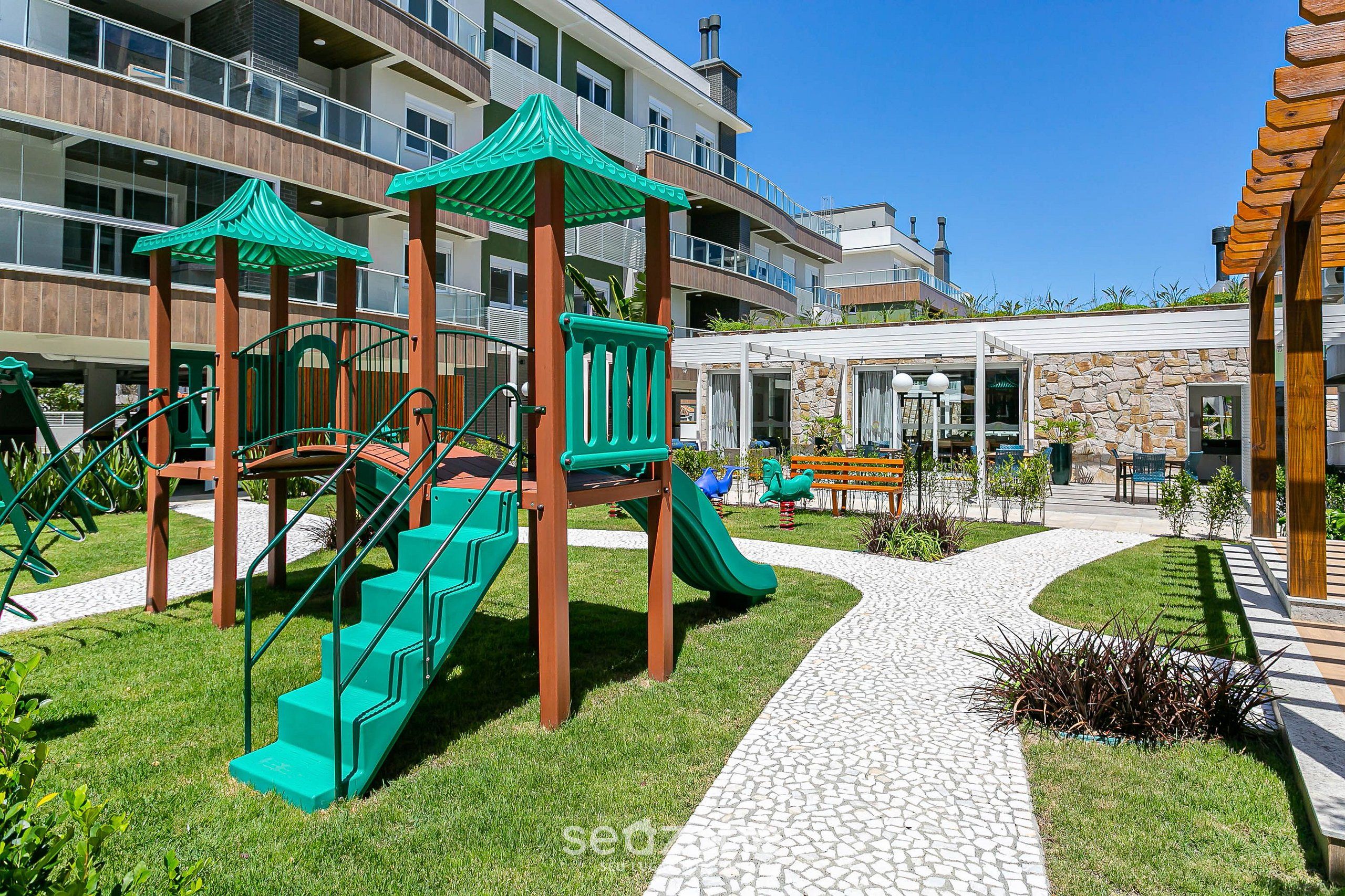Un parque infantil con toboganes verdes y escaleras, rodeado de un camino pavimentado y vegetación.