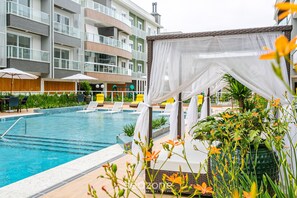A swimming pool with lounge chairs and umbrellas, surrounded by a multi-story building with balconies. - Seaside Condominium in Campeche-florianopolis SHR (Florianópolis)