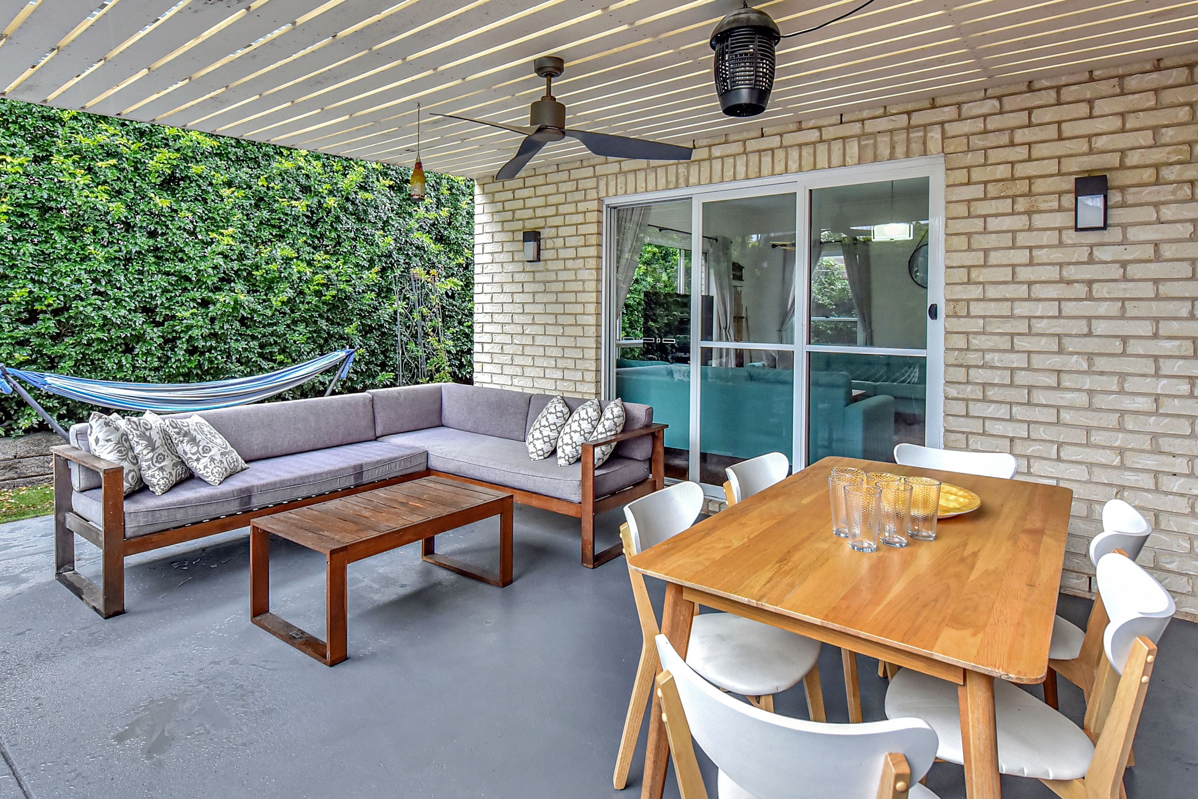 Une terrasse extérieure avec une table en bois, des chaises blanches, un canapé gris agrémenté de coussins à motifs, un hamac et un mur de briques surmonté d’une haie verte.