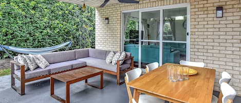 Un patio al aire libre con una mesa de madera, sillas blancas, un sofá gris con cojines estampados, una hamaca y un muro de ladrillo con un seto verde.