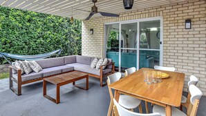 An outdoor patio with a wooden table and chairs, a grey sofa with patterned cushions, a hammock, and a brick wall with a green hedge.