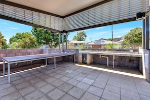 Área externa com assentos, um banco comprido e uma mesa, cercada por um teto alto com ripas horizontais, piso de azulejos e vista para edifícios residenciais e árvores.