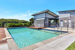 Una piscina con cubierta de madera, un espacio de estar techado y un edificio de fachada gris.