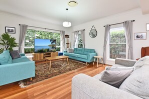 Ein Wohnzimmer mit einer Couch, einem Couchtisch und einem Fernseher. Es gibt Fenster mit Vorhängen, eine Pflanze und einen Spiegel an der Wand.