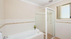 Ein Badezimmer mit einer gläsernen Duschkabine, einer Badewanne mit Handtuch und Kerze sowie einem Fenster mit Jalousien.