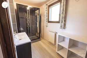 A bathroom with a glass-enclosed shower, a white sink, and a window with patterned curtains. - Chalet T6 confort proche Station (Dévoluy)