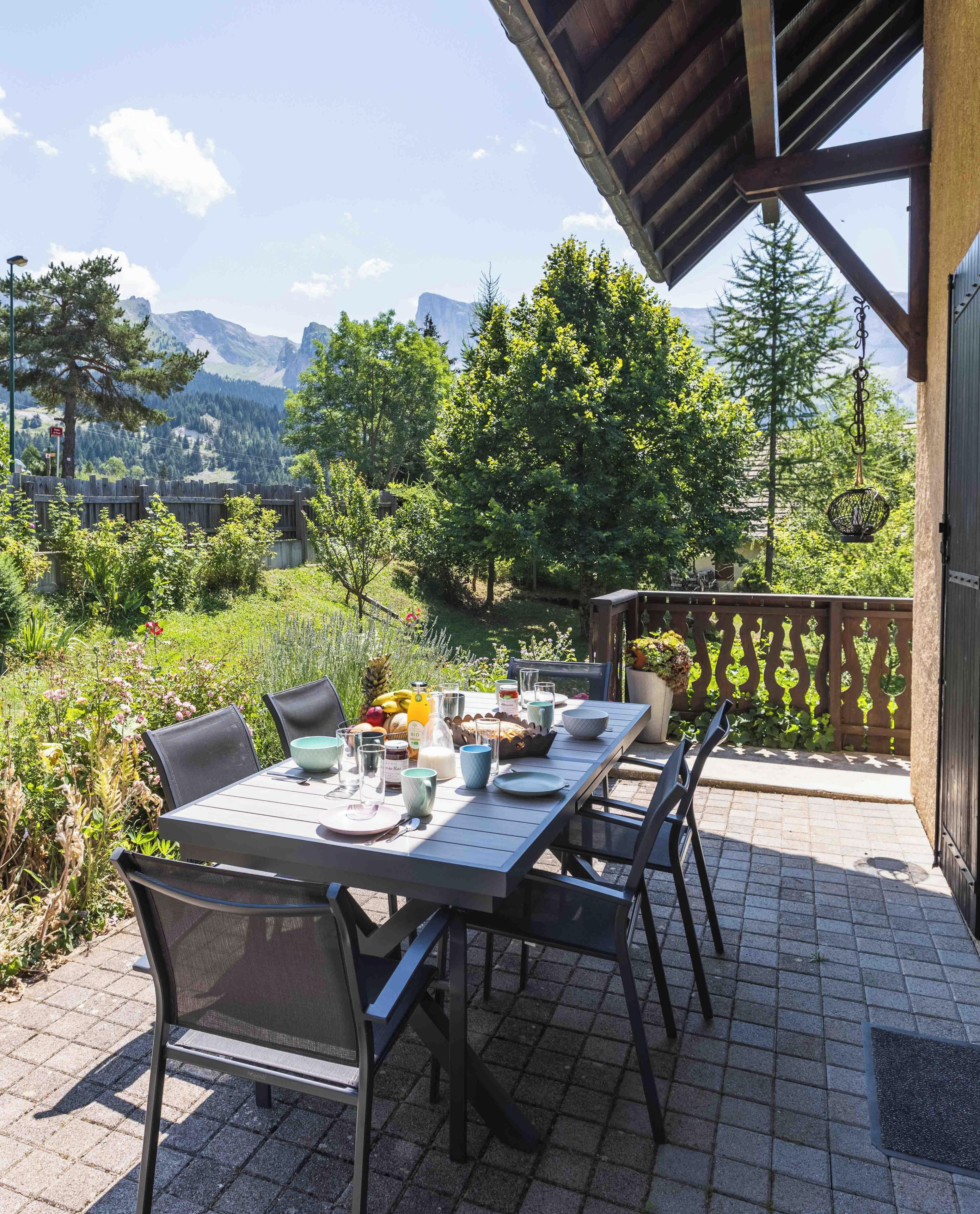 An outdoor dining area with a table set for two, surrounded by chairs, with a view of mountains and trees in the background.
