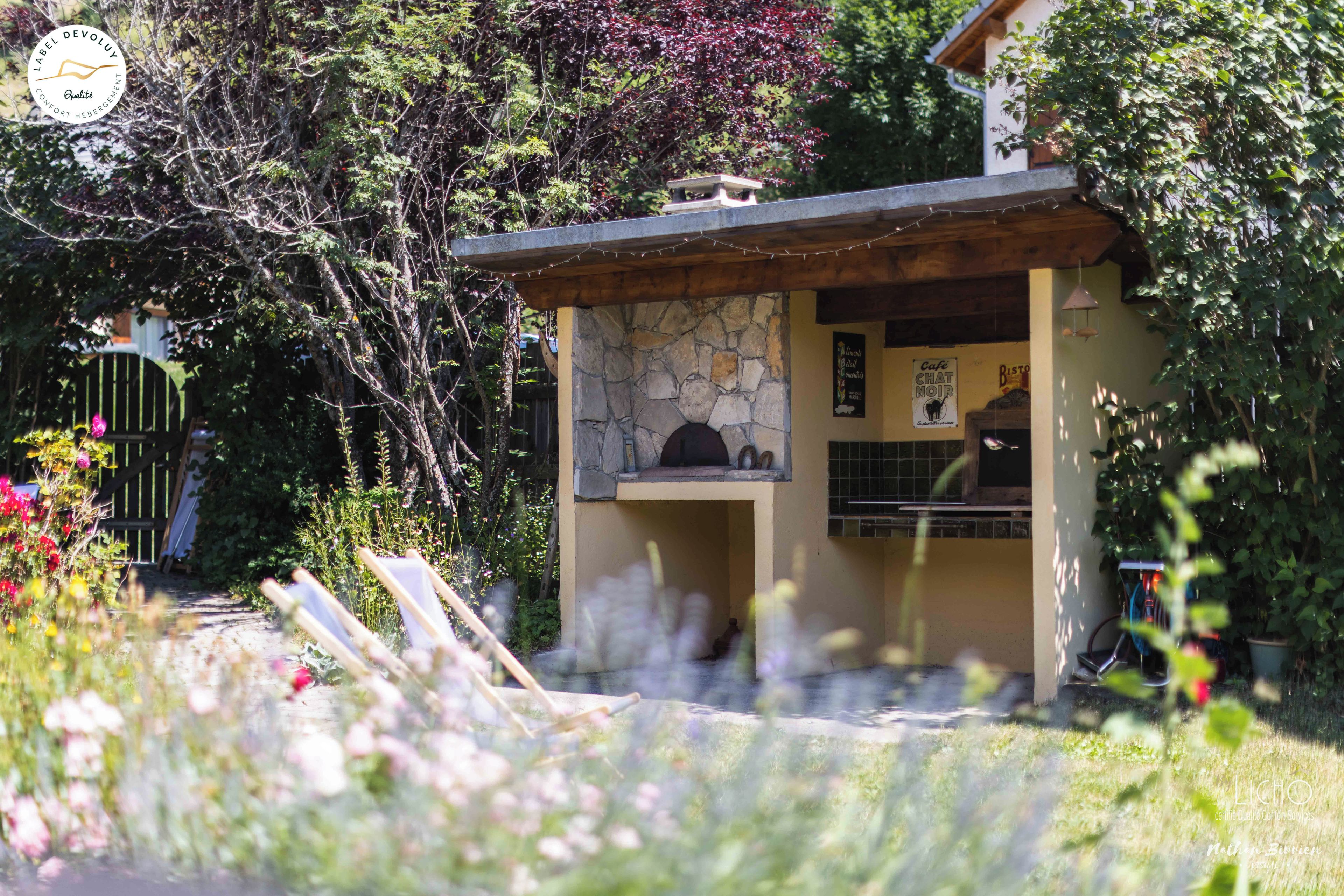 Un edificio in pietra con un piccolo portico, immerso nel verde e con un vialetto fiorito.