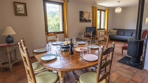 A dining area with a wooden table set for four, surrounded by wicker chairs with cushions. A television is mounted on the wall, and a wood-burning stove is visible in the background. - Chalet T6 confort proche Station (Dévoluy)