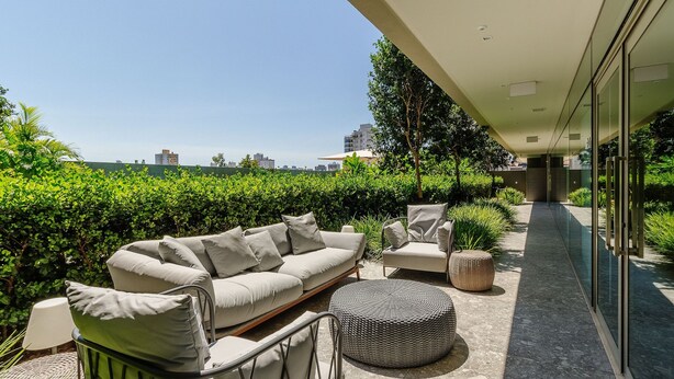 Een moderne buitenruimte met een bank en stoelen, een rond poef, en een glazen balustrade met uitzicht op een verzorgde tuin en stadsbeeld.