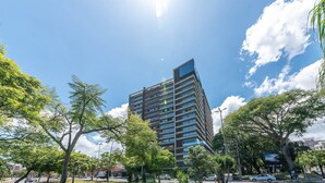 Een modern hoog gebouw met meerdere verdiepingen, omringd door weelderige groene bomen en een heldere blauwe lucht.