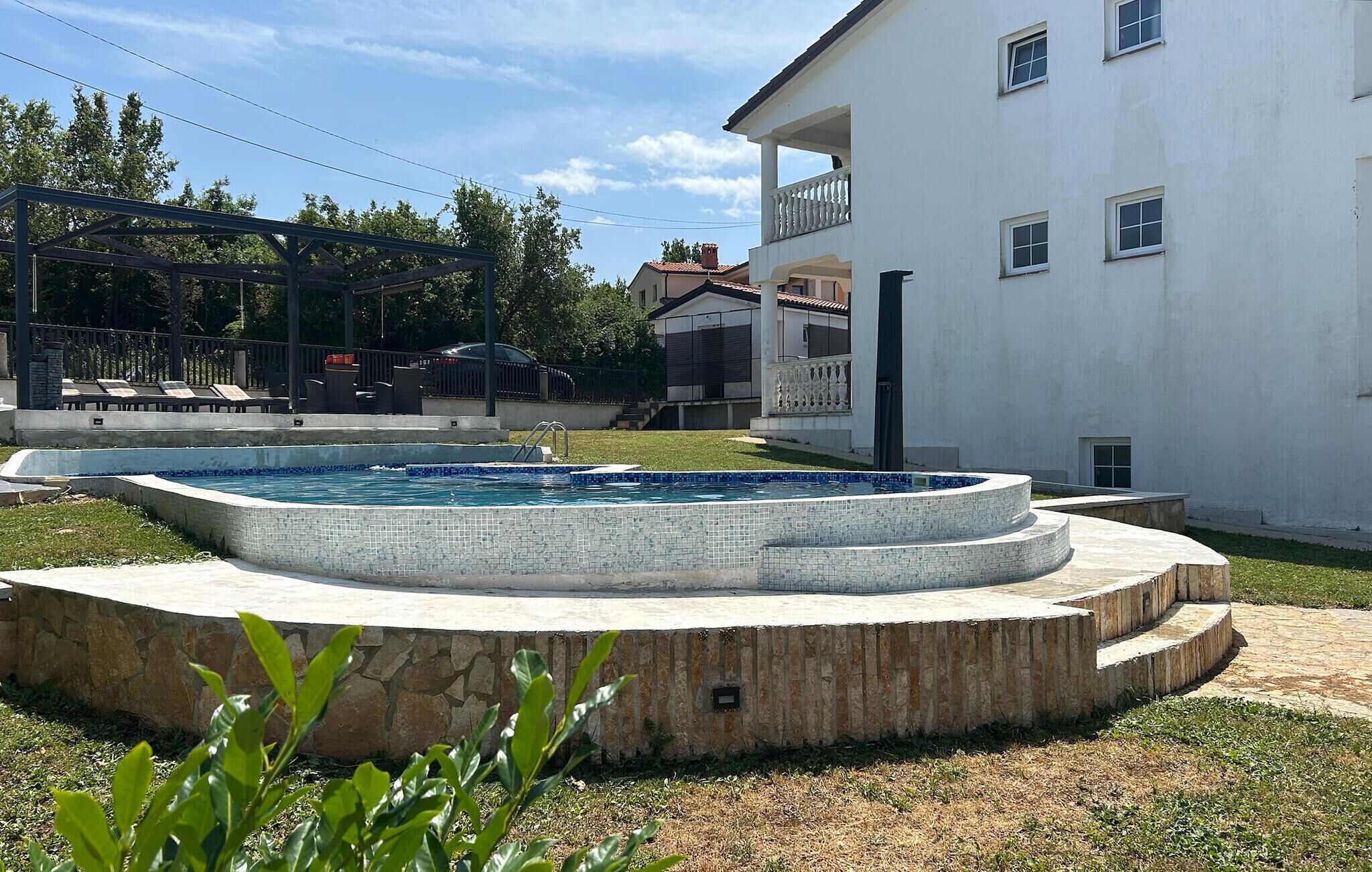 A swimming pool with a tiled edge, surrounded by a stone deck. There is a building with a balcony in the background.
