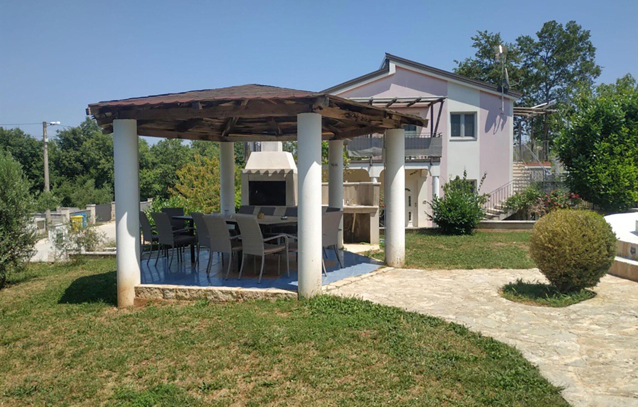 A covered outdoor dining area with a grill, surrounded by a stone pathway and greenery.