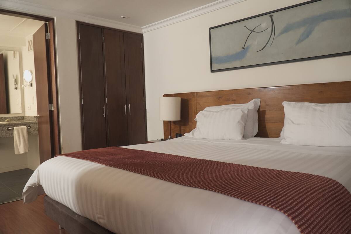 Une chambre d’hôtel avec un grand lit, une tête de lit en bois, du linge de lit blanc, un plaid rouge, une table de chevet avec une lampe, une armoire en bois et un tableau abstrait encadré accroché au mur.
