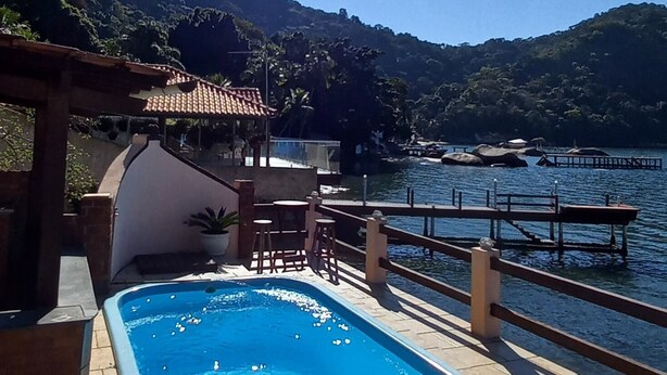 A swimming pool with a wooden deck and a pergola, surrounded by a scenic view of hills and a body of water. - Excellent house on Águas Lindas beach (Itacuruça Island) (Itacuruçá Mangaratiba)