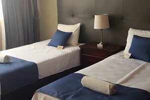 Two single beds with blue and white bedding, a nightstand with a lamp, and a grey headboard.