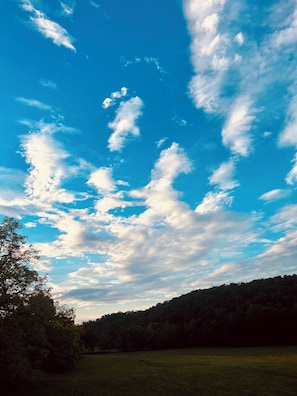 Cielo despejado con algunas nubes dispersas sobre un campo cubierto de hierba y una colina boscosa.