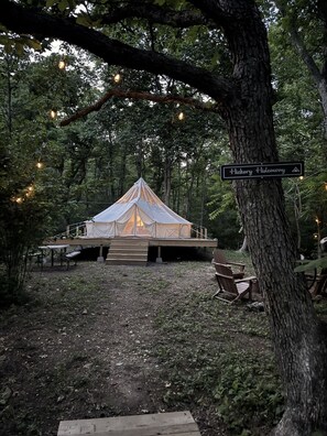 A campsite with a tent, wooden deck, and seating area, surrounded by trees and string lights.